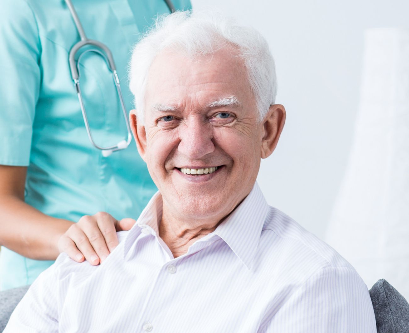Ein älterer Mann mit einem Lächeln sitzt auf einer Couch, begleitet von einer fürsorglichen Pflegerin.