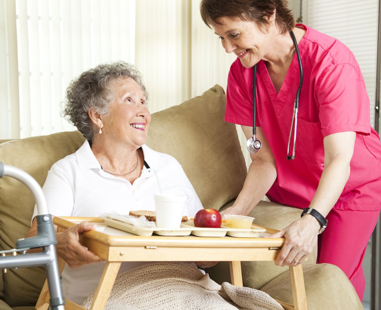 Eine Pflegekraft unterstützt eine ältere Dame, während sie ein Tablett mit Speisen oder Getränken reicht.