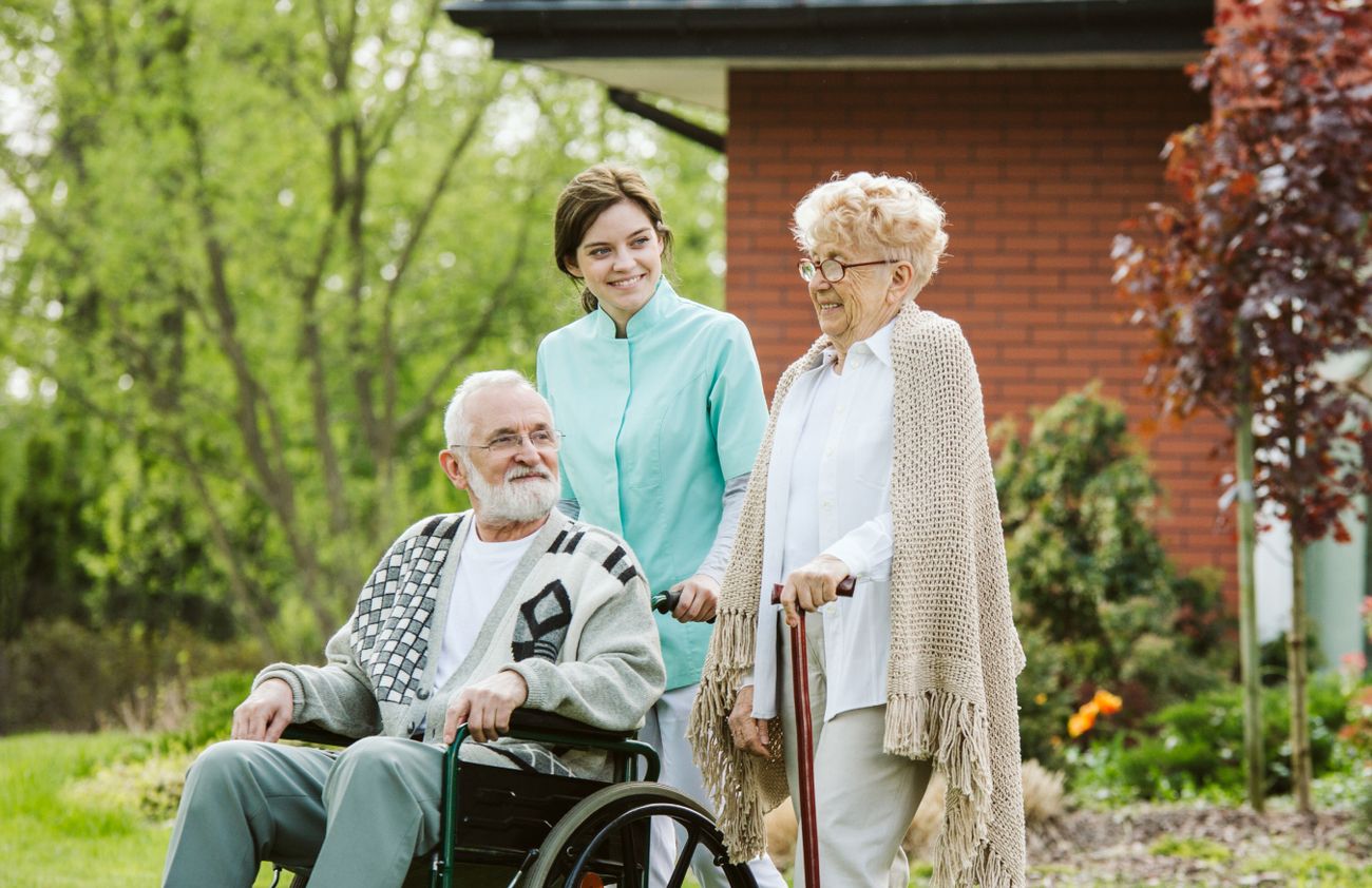 Ein älterer Mann sitzt im Rollstuhl, während zwei Frauen ihm Unterstützung und Gesellschaft bieten.
