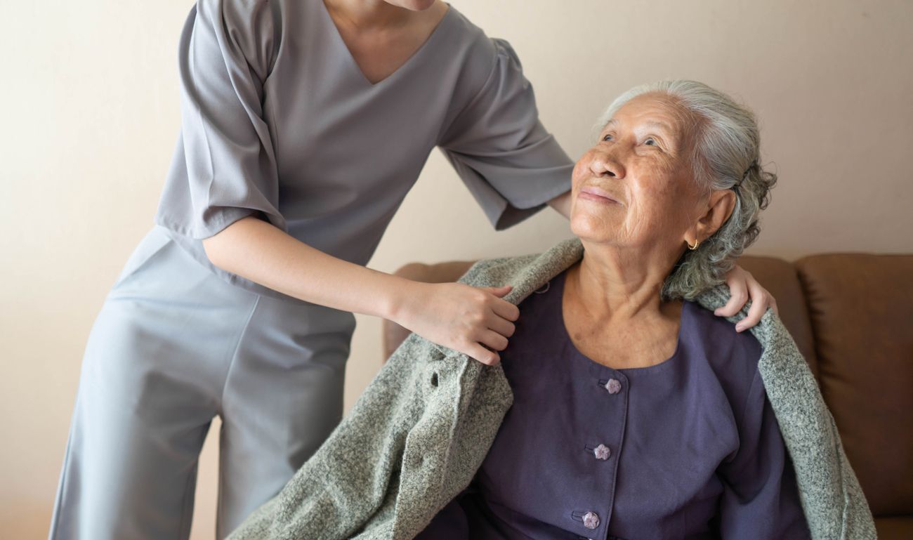 Eine junge Frau unterstützt eine ältere Dame auf einem Sofa, indem sie ihr hilft und ihr zur Seite steht.