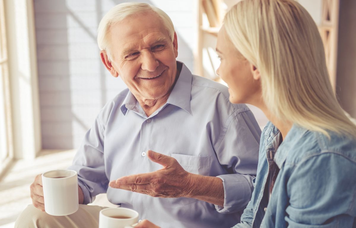 Auf einer Couch sitzen ein älterer Mann und eine ältere Frau, beide halten Kaffeetassen und wirken entspannt und zufrieden.